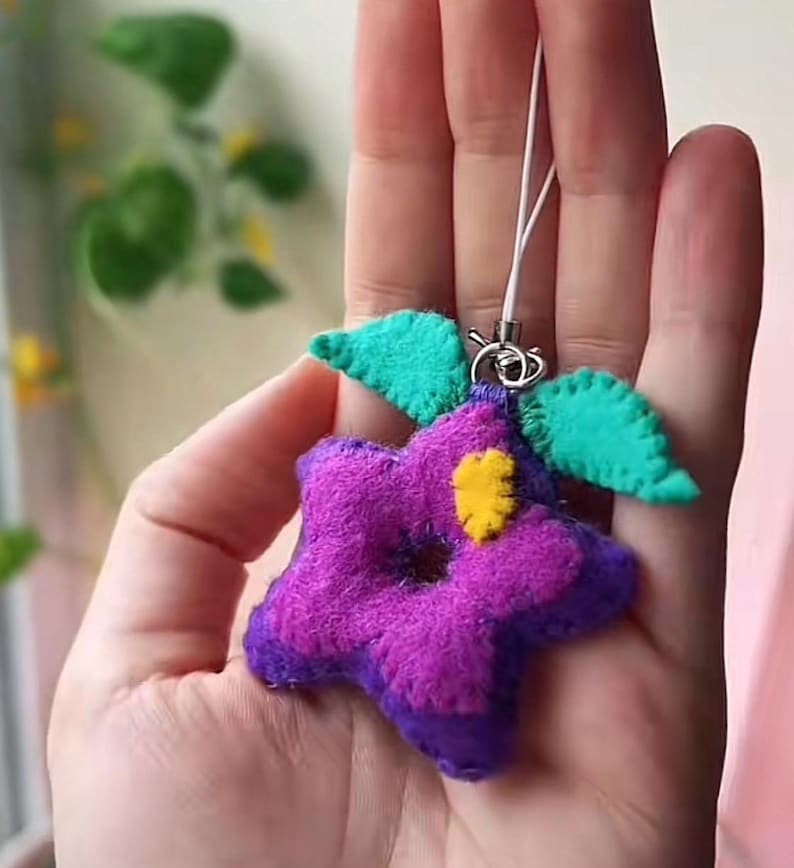 Pu&ograve; includere: Un fiore di feltro viola e blu con un centro giallo e foglie di feltro verde attaccate a una catena di ciondolo per telefono bianca.