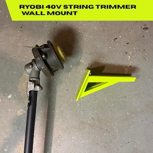 以下が含まれることがあります： Ryobi 40Vストリングトリマー用のネオングリーン壁掛けブラケット。ストリングトリマーの黒いシャフトとカッティングヘッドの横に配置されています。画像上部には黒字でRYOBI 40V STRING TRIMMER WALL MOUNTというテキストが表示されています。