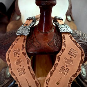 May include: A pair of light tan leather spur straps featuring intricate floral tooling along the edges and the embossed letters E.R. with a small bucking horse graphic in the center. The straps are attached to silver-toned metal buckles.