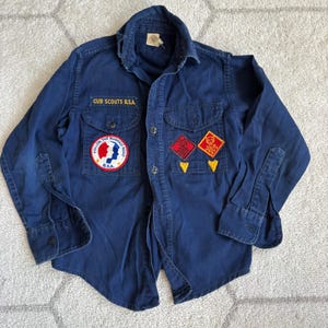 May include: A vintage navy blue Cub Scouts B.S.A. shirt. The shirt features two chest pockets and several patches, including one that reads "Follow the Rugged Road." The text "Cub Scouts B.S.A." is embroidered above the left pocket.
