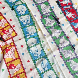 Könnte beinhalten: Weißer Stoff mit Bildern von Zeichentrickfiguren in farbenfrohen Rahmen. Das Design umfasst Tweety, Bugs Bunny und andere Looney Tunes-Charaktere. Der Hintergrund hat verstreute Dreiecke in verschiedenen Farben.