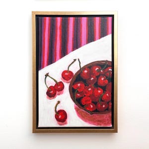 May include: A framed painting shows a still life of cherries. A brown bowl is filled with bright red cherries, with a few scattered on a white surface. The background features pink and purple vertical stripes. The painting is in a gold frame.