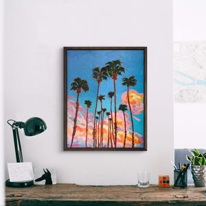 May include: An oil painting of palm trees against a blue sky with pink and orange clouds. The artwork is framed in a dark brown wooden frame. The painting depicts tall, slender palm trees with green fronds.