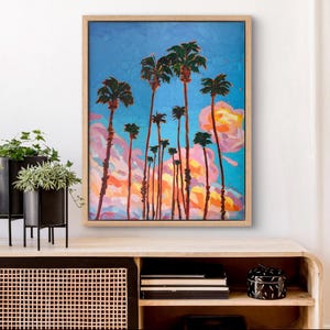 May include: An oil painting of palm trees against a vibrant blue sky with pink and orange clouds. The artwork is framed in a light wood frame and evokes a tropical landscape. The painting is displayed on a wooden cabinet.