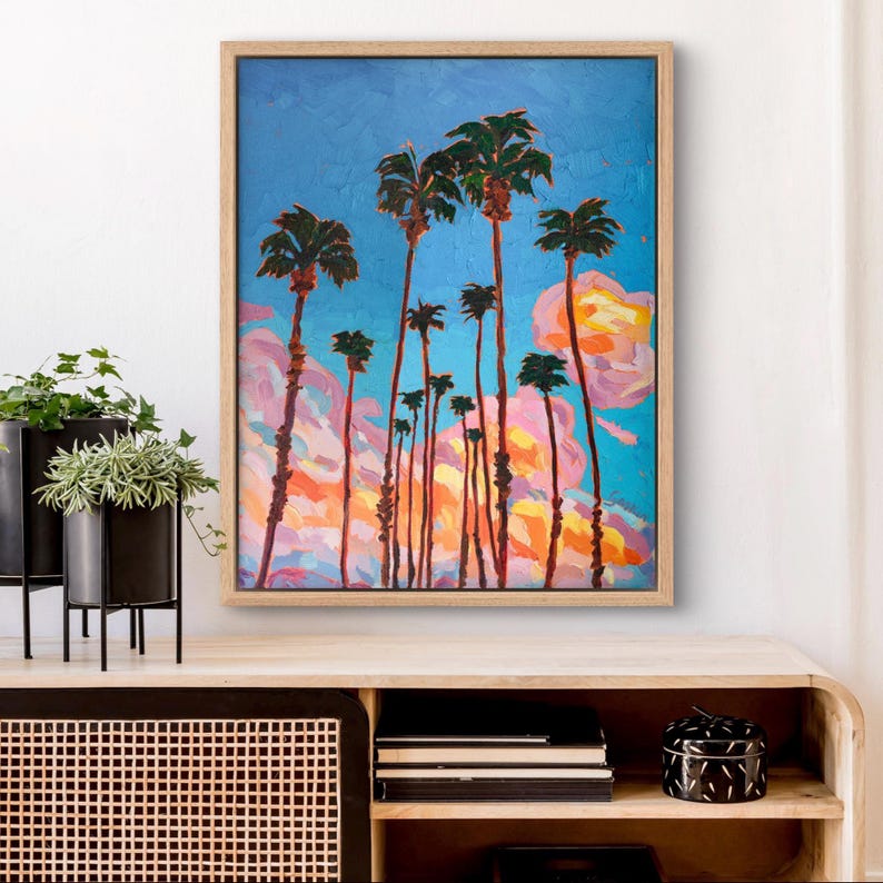 May include: An oil painting of palm trees against a vibrant blue sky with pink and orange clouds. The artwork is framed in a light wood frame and evokes a tropical landscape. The painting is displayed on a wooden cabinet.