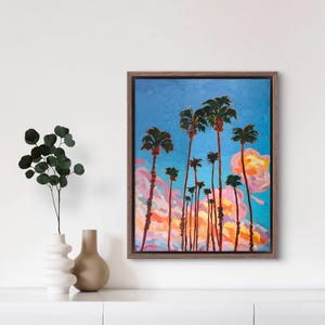 May include: An oil painting of palm trees against a blue sky with pink and orange clouds. The artwork is framed in a brown wooden frame. The painting has a textured, impressionistic style.