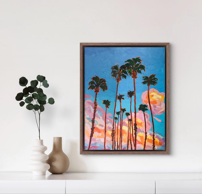 May include: An oil painting of palm trees against a blue sky with pink and orange clouds. The artwork is framed in a brown wooden frame. The painting has a textured, impressionistic style.