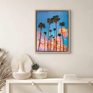 May include: An oil painting of palm trees against a blue sky with pink and orange clouds. The artwork is framed in a light wood frame. The painting depicts a tropical landscape.