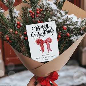 May include: A Christmas bouquet featuring evergreen branches, red berries, and a card. The card reads "Merry Christmas" with a red bow illustration and the text "With Love, The Young Family, 2030". The bouquet is wrapped in brown paper.
