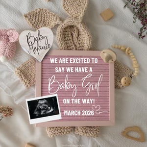 Peut inclure: Un tableau rose annonce "Baby Girl" avec le texte "Nous sommes ravis de vous annoncer que nous attendons une petite fille ! Mars 2026." Une photo d'échographie et un cœur en bois avec le nom "Melanie" sont inclus. Des oreilles de lapin tricotées et une sucette sont également présentes.