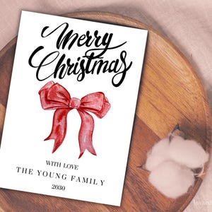 May include: A white Christmas card with the words "Merry Christmas" in elegant black script. A watercolor-style red bow is centered below the text. The card includes the text "With Love, The Young Family, 2030".