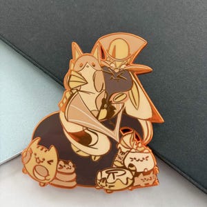 May include: A gold-colored enamel pin featuring a cartoon character with a long hat and cape, surrounded by several small cat-like figures. The pin has a brown and tan color scheme with black accents.