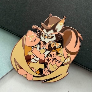 May include: A detailed enamel pin featuring a cartoon cat character with a hat, bow tie, and wings. The pin has a color palette of brown, tan, and peach. The cat is surrounded by smaller cartoon characters. The pin is on a gray surface.