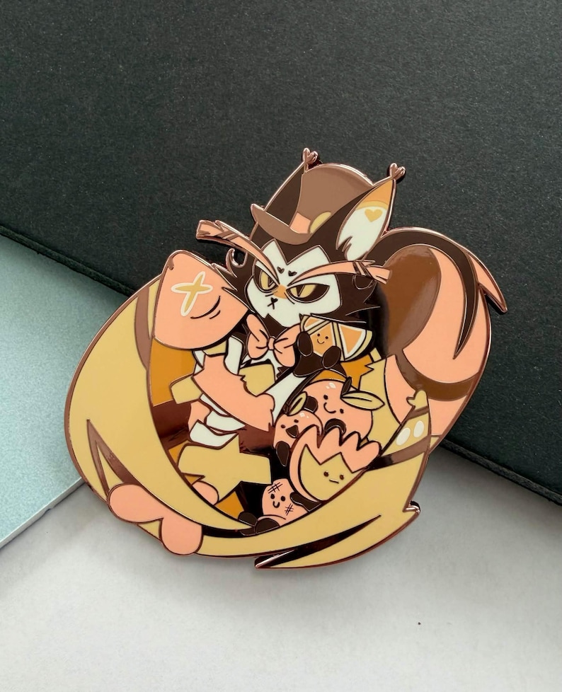 May include: A detailed enamel pin featuring a cartoon cat character with a hat, bow tie, and wings. The pin has a color palette of brown, tan, and peach. The cat is surrounded by smaller cartoon characters. The pin is on a gray surface.
