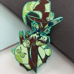 May include: A green and brown enamel pin featuring a cartoon witch character with a hat and a skull. The pin has a shiny, reflective finish and includes two small animal characters. The overall design is whimsical and detailed.