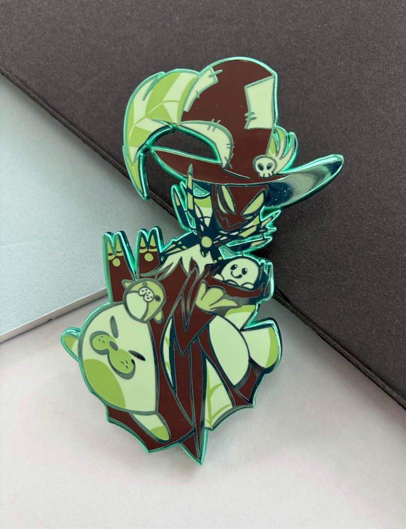 May include: A green and brown enamel pin featuring a cartoon witch character with a hat and a skull. The pin has a shiny, reflective finish and includes two small animal characters. The overall design is whimsical and detailed.