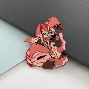 May include: A red and pink enamel pin featuring a cartoon character with a hat, wings, and a snake-like detail. The pin has brown accents and a detailed design. The pin is on a gray and white surface.