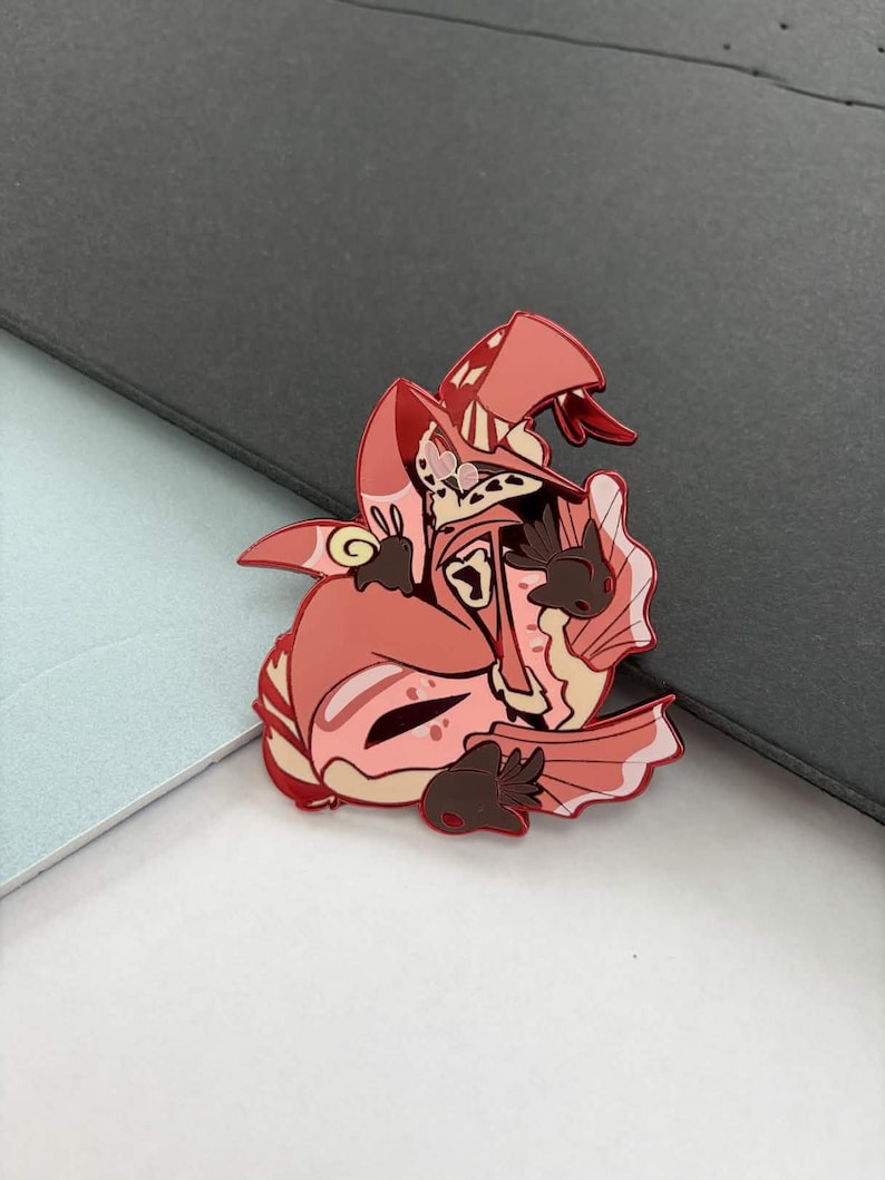 May include: A red and pink enamel pin featuring a cartoon character with a hat, wings, and a snake-like detail. The pin has brown accents and a detailed design. The pin is on a gray and white surface.