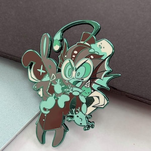 May include: A teal, brown, and white enamel pin featuring a cartoonish creature with large eyes, a rabbit, and a small frog. The pin has a shiny, metallic finish and is set against a dark background. The design is whimsical and detailed.