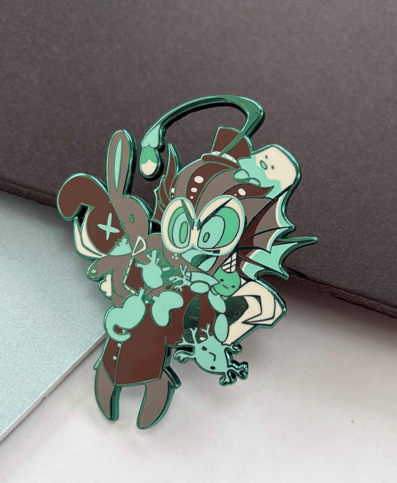 May include: A teal, brown, and white enamel pin featuring a cartoonish creature with large eyes, a rabbit, and a small frog. The pin has a shiny, metallic finish and is set against a dark background. The design is whimsical and detailed.