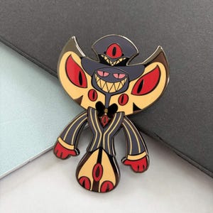 May include: A colorful enamel pin featuring a stylized demon character with a dark gray hat, yellow wings, and a black and yellow striped suit. The pin has red eyes and a wide, toothy grin.