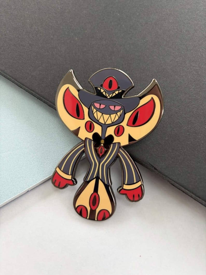 May include: A colorful enamel pin featuring a stylized demon character with a dark gray hat, yellow wings, and a black and yellow striped suit. The pin has red eyes and a wide, toothy grin.