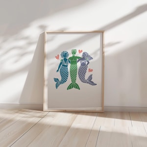 Könnte beinhalten: Eine gerahmte Illustration von drei Meerjungfrauen, zwei Frauen und ein Mann, mit grünen, blauen und violetten Schwänzen. Sie halten sich an den Händen und lächeln. Um sie herum befinden sich kleine rote Herzen.