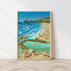 Op de afbeelding: Een ingelijste kunstdruk die een kustscène met een strand, oceaan en mensen weergeeft. Het kunstwerk toont een blauwe lucht, turquoise water en een zandstrand. Surfers rijden op golven en mensen ontspannen op de kust. De print zit in een houten lijst.