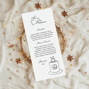 May include: A white menu card with a black and white illustration of a teddy bear on top of a cake. The menu card lists the starter, main course, and dessert for a baby shower. The text on the menu card reads "Starter", "Caprese Salad with Balsamic Glaze", "Main Course", "Herb-Crusted Chicken with Garlic Mashed Potatoes & Seasonal Vegetables", "Dessert", "Cake", and "Oh Baby".