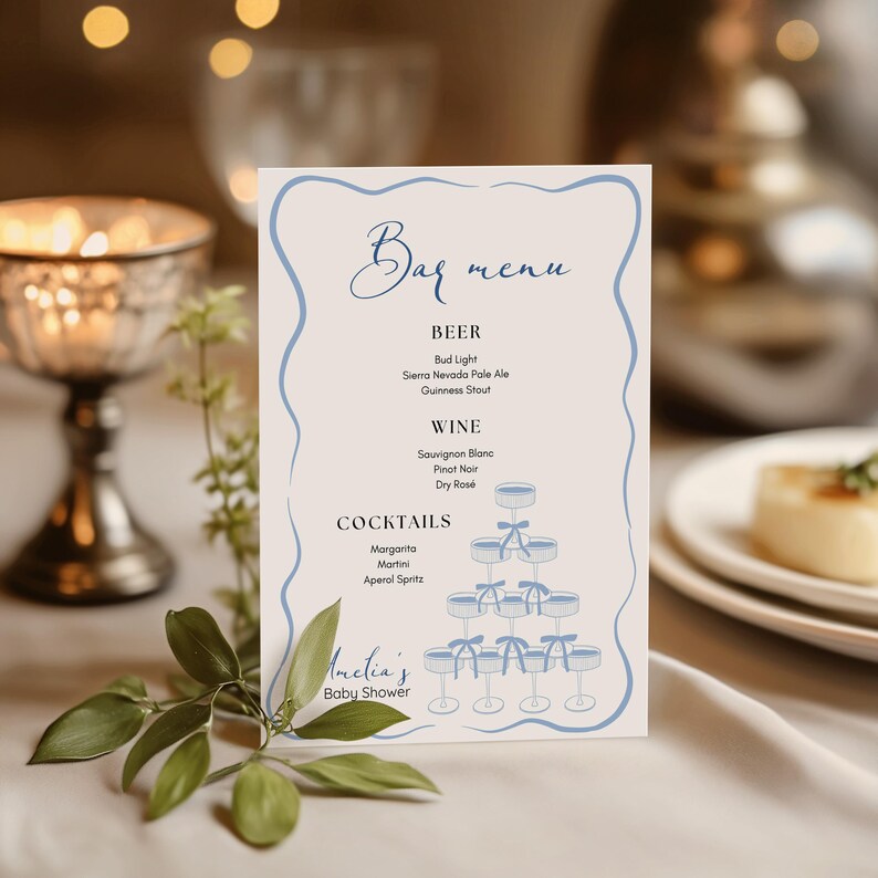 May include: A white bar menu with blue accents and a blue illustration of a tiered cake stand with glasses. The menu lists beer, wine, and cocktails. The text at the bottom reads "Amelia's Baby Shower".