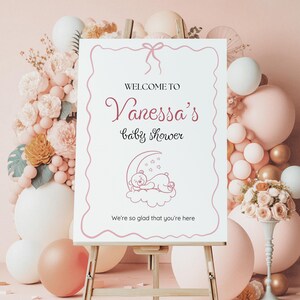 May include: A white sign with pink text and a pink ribbon on top. The sign says "Welcome to Vanessa's Baby Shower" and has a drawing of a teddy bear sleeping on a crescent moon. The text at the bottom of the sign says "We're so glad that you're here."