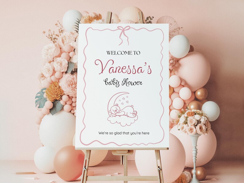 May include: A white sign with pink text and a pink ribbon on top. The sign says "Welcome to Vanessa's Baby Shower" and has a drawing of a teddy bear sleeping on a crescent moon. The text at the bottom of the sign says "We're so glad that you're here."