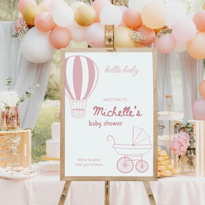 May include: A pink and white baby shower sign with a hot air balloon and a baby carriage. The sign says "hello baby" and "Welcome to Michelle's baby shower".