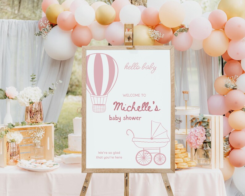 May include: A pink and white baby shower sign with a hot air balloon and a baby carriage. The sign says "hello baby" and "Welcome to Michelle's baby shower".