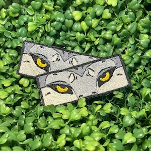 May include: Two embroidered patches featuring a stylized wolf eye design. The patches have a gray background with yellow eyes and black outlines. The patches are rectangular and are set against a backdrop of green foliage.