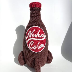May include: Crocheted brown bottle with a red cap and a white and red "Nuka Cola" logo. The bottle has two small, brown, rocket-like protrusions at the base. The handmade item is likely a decorative piece or a toy.