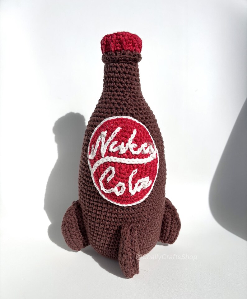 May include: Crocheted brown bottle with a red cap and a white and red "Nuka Cola" logo. The bottle has two small, brown, rocket-like protrusions at the base. The handmade item is likely a decorative piece or a toy.