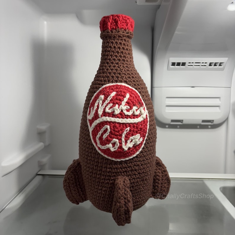 May include: A brown crocheted bottle with a red top and a red and white circular emblem that reads "Nuka Cola". The bottle has two brown fins at the bottom. The bottle is inside a refrigerator.