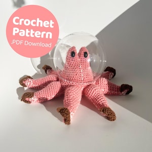 Puede incluir: Un pulpo de crochet rosa con detalles marrones se encuentra dentro de una cúpula de vidrio transparente. El pulpo tiene un diseño de crochet blanco en la cabeza. La imagen incluye el texto "Crochet Pattern .PDF Download".
