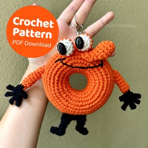 May include: An orange crocheted donut-shaped keychain with black and white details, featuring arms, legs, and large eyes. The keychain has a smiling face and the text "Crochet Pattern .PDF Download" in an orange circle.