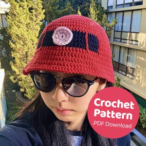 Puede incluir: Sombrero de crochet rojo con un diseño negro y rosa. El sombrero tiene un ala ancha y está tejido en punto texturizado. El texto "Crochet Pattern .PDF Download" está en un círculo rojo en la esquina inferior derecha de la imagen.