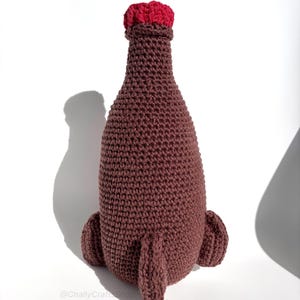 May include: A handmade, crocheted bottle in shades of brown with a red top. The bottle has a rounded body and two small, rounded protrusions at the base. The crochet work is visible, giving the bottle a textured appearance. The bottle is displayed on a white background.