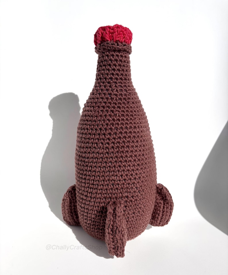 May include: A handmade, crocheted bottle in shades of brown with a red top. The bottle has a rounded body and two small, rounded protrusions at the base. The crochet work is visible, giving the bottle a textured appearance. The bottle is displayed on a white background.