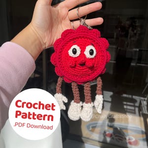 Puede incluir: Un llavero rojo tejido a ganchillo con forma de personaje de dibujos animados con ojos blancos, una cara sonriente y piernas marrones y blancas. El llavero tiene un anillo plateado. El texto "Crochet Pattern .PDF Download" está en la esquina inferior izquierda.