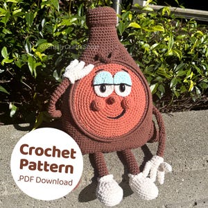 Peut inclure: Jouet amigurumi crocheté en forme de bouteille brune avec un visage rouge et des yeux blancs. Le jouet a des bras et des jambes bruns et des pieds blancs. Le jouet est assis sur une surface grise. Le texte "Crochet Pattern .PDF Download" est sur un cercle blanc.