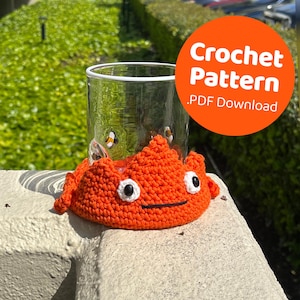 Puede incluir: Posavasos de ganchillo naranja con una cara sonriente y ojos negros. El posavasos está sobre una superficie gris con una taza de vidrio dentro. El texto "Crochet Pattern .PDF Download" está en un círculo naranja brillante.