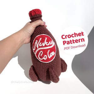 May include: A brown crochet bottle with a red cap and a red and white circular label that reads "Nuka Cola". The bottle is held by a hand. A white circle with the text "Crochet Pattern .PDF Download" is in the background.