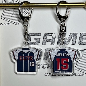 May include: Two baseball jersey keychains with clear acrylic backing. The jersey on the left is navy blue with white pinstripes and red lettering that says "ELITE 15". The jersey on the right is navy blue with white pinstripes and red lettering that says "MELTON 15".