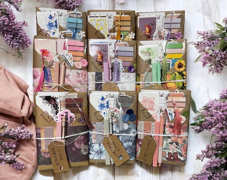 May include: A collection of wrapped gift sets, each featuring a variety of stationery items. The sets include notebooks, pens, bookmarks, and decorative elements like tassels and floral accents. The color palette is soft, with pinks, browns, and floral patterns.