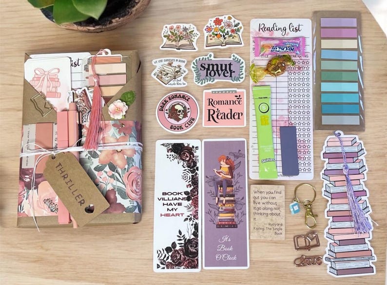 May include: A collection of book-themed items, including stickers, bookmarks, and a gift box. The stickers feature open books with floral designs, and the bookmarks have quotes and illustrations. The gift box is wrapped in floral paper and tied with a tassel.